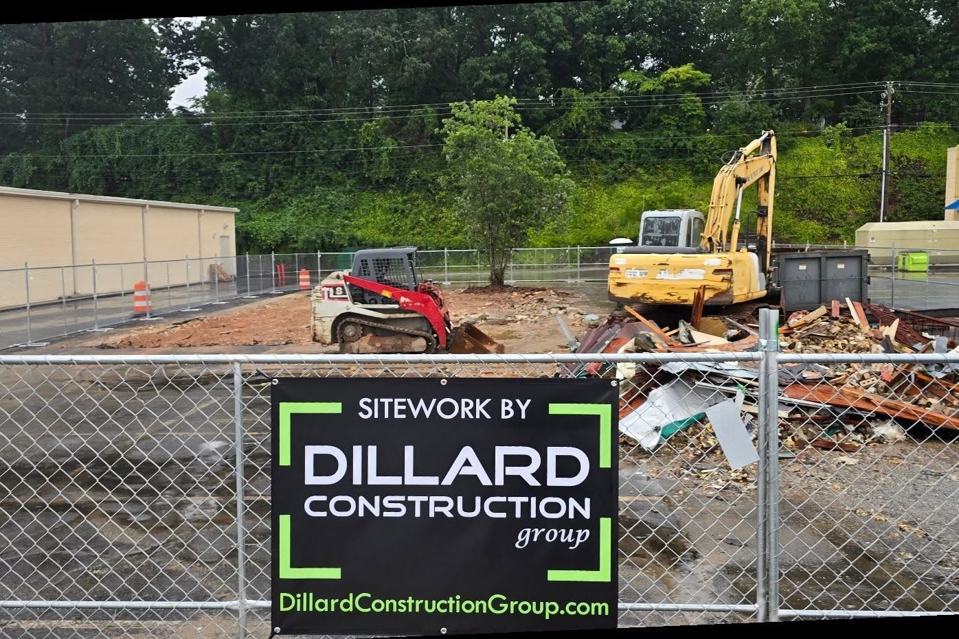 A Sitework by Dillard Construction Group sign on the fence, with a skid steer, an excavator and demolition debris behind it in Easley, SC.