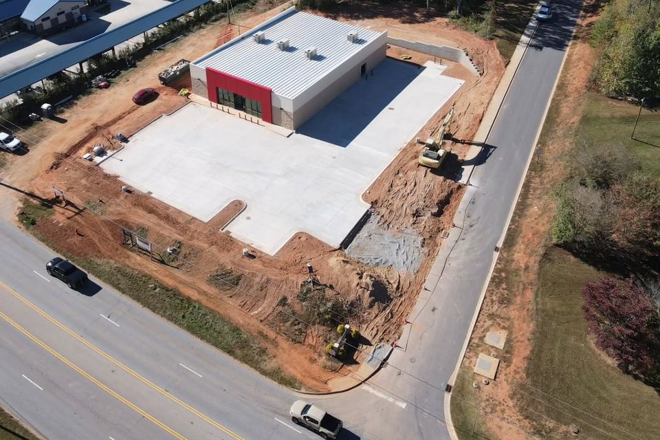 Aerial view of the Pickens County retail pad: the new building, its fresh concrete lot, graded slopes and an excavator beside the road.