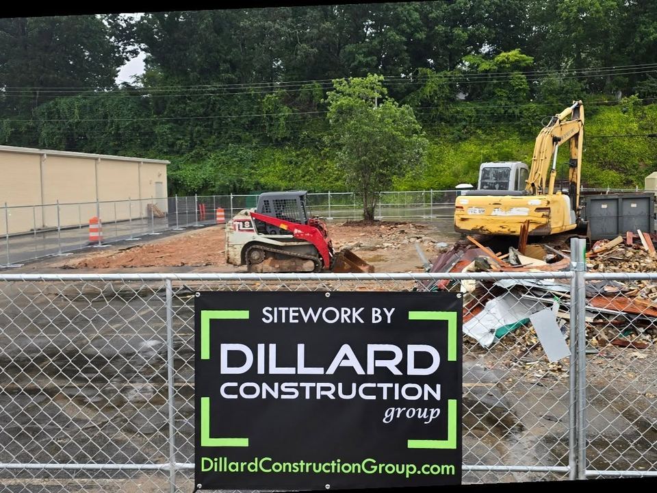 A Sitework by Dillard Construction Group sign on the fence in Easley, SC, with demolition debris, a skid steer and an excavator behind it.