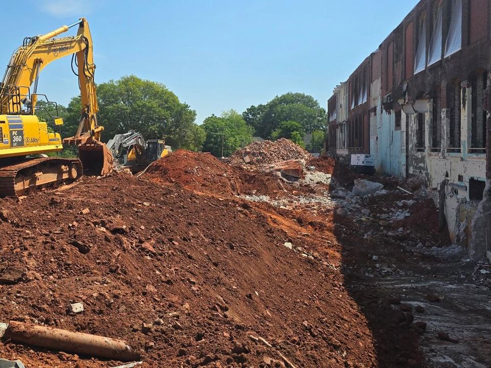 Excavator digging beside the brick wall of a historic textile mill in Greenville, SC during site work.